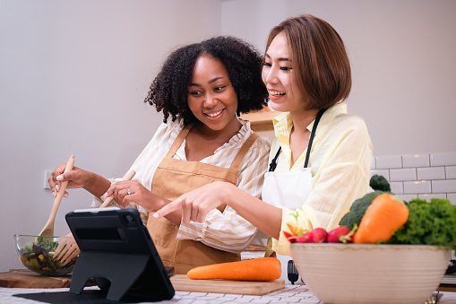 Beautiful homosexual Lesbian couples learning to cooking healthy meals,African American Asian woman LGBTQ+,LGBT,LGBTQ home online kitchen.Wellness lifestyle.Tablet learning.Salad Diet foods .