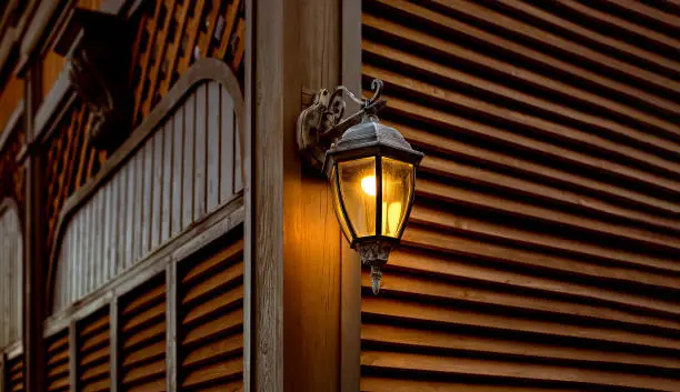 iron retro lantern with a glass shade and electric light bulb, object mounted on wooden wall and shutters made of planks, architecture of evening lighting with a warm glow.