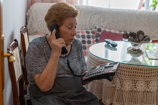 Elderly woman speaks on the phone, sitting on a chair inside her house