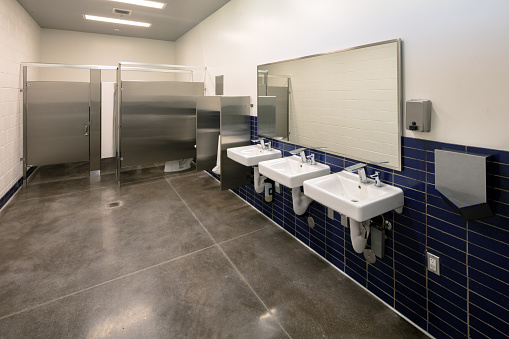 Modern public bathroom for boys. Gray and stainless steel. Boys bathroom with urinal and toilet stalls. White Sinks with large mirror