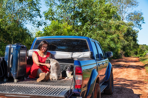 A woman sitting in the back of a pickup truck with her dogs and suitcase, all on a typical red sand road in Paraguay.