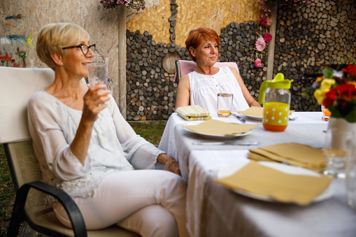 Selective focus shot of two senior women enjoying drinks and listening to their friends sharing interesting stories during a dinner party in the back yard.