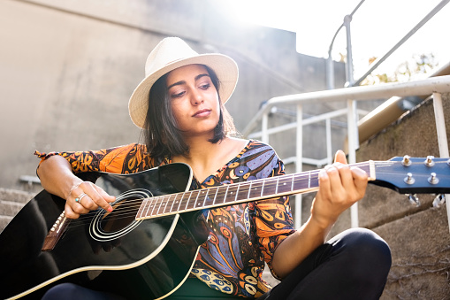 Portrait of a beautiful young woman musician wearing a hat playing an acoustic guitar while sitting outside on steps