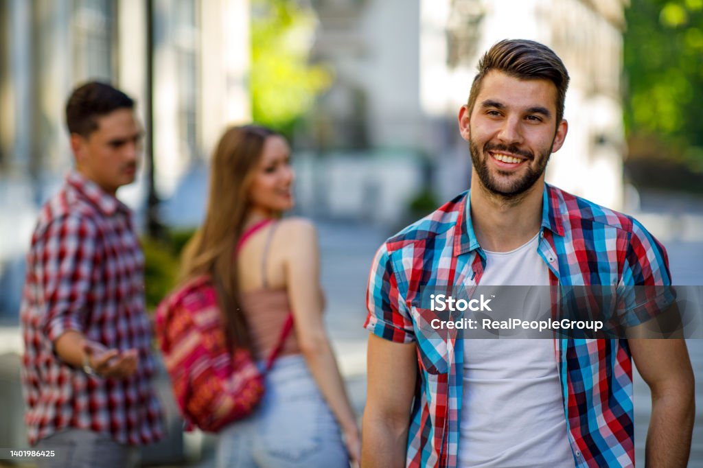 Young woman makes a jealous situation while walking around the city with her boyfriend Disloyal young woman looking another handsome man and her angry boyfriend looking at her on the street Envy Stock Photo