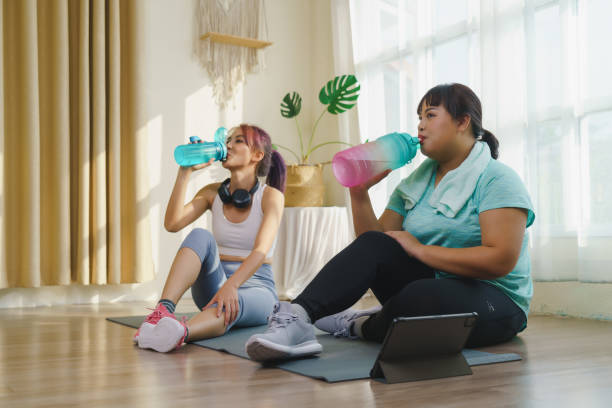 istock Thirst while exercising concept. Two Asian women's body size is different in sportswear, drinking water while resting and sitting on yoga mat after fitness exercise at home together. 1401991055