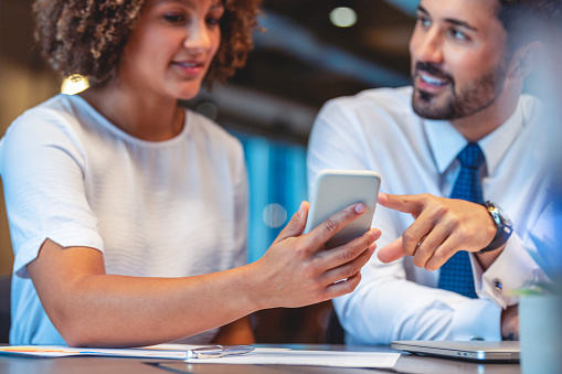 Business man and woman working and looking at a mobile phone in the office. They are both wearing business clothes and are looking happy. There is paperwork and technology on the table.