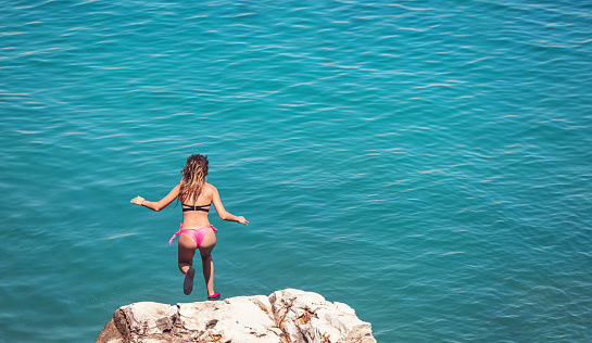 Young girl having fun at the beach on summer vacations in Greece. She jumping from cliff and diving into blue sea