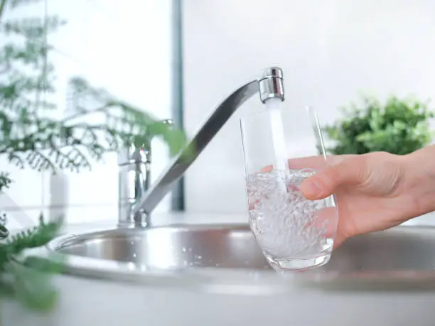 Photo of A stream of clean water drink flows into the glass. Woman holding a glass of water under running water from the tap in the kitchen