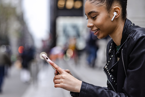 Side view of young African American woman in the city using mobile phone. She is using airpods and smiling.