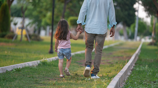 Photo of father holding 4 years old daughter's hand and walking in public park. Selective focus on models. Shot in outdoor with a full frame mirrorless camera.