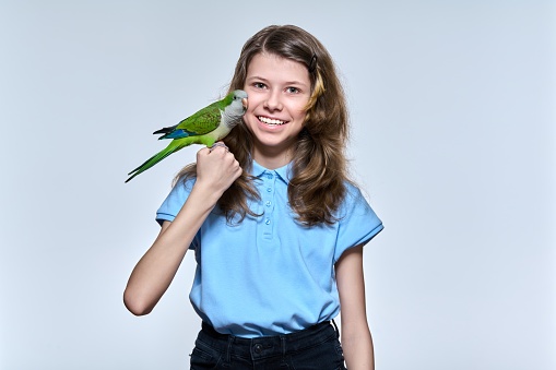 Smiling child girl with pet green quaker parrot looking at camera on light studio background. Animals, bird owner, childhood, child and pet friendship concept