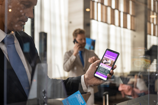Businessman seen holding his phone with a booking app on the screen while his colleague is talking on the phone just before they are checking in during a business trip.