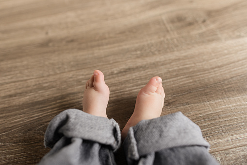 Leg & Feet of a 24-Week-Old Baby Boy With 12 Toes Wearing a Blue Denim Jeans While Sitting on a Wooden Floor in a Bright Home.