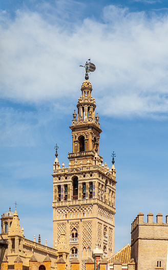 Seville, Spain - 8th April 2022. Detail of the top section of La Giralda belltower in Sevilla, seen over the rooftops. The lower part of the tower was originally the minaret of a mosque and was completed in 1198. The belfry was completed c1568, with the addition of the Giraldilla (weather vane). The Giralda is a UNESCO World Heritage Site.