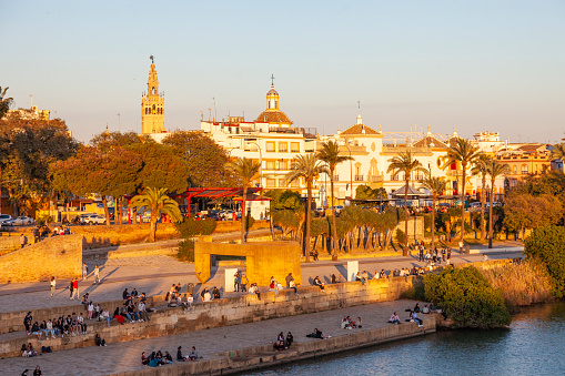 Seville, Spain - 7th April 2022.  Seville, Spain - 7th April 2022 People, mostly young people, enjoying a summer evening on the Muelle de la Sal area of the Paseo Alcalde Marques del Contadero, beside the Canal de Alfonso XIII, part of the Guadalquivir river.  The Monument to Tolerance (1992) by Eduardo Chillida is near the centre, and La Giralda can be seen behind.
