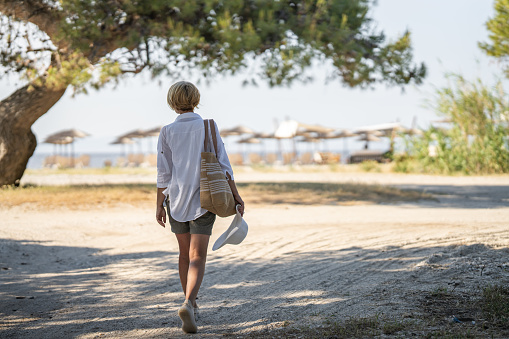 Mature woman carrying a bag and a hat is walking towards the beach.