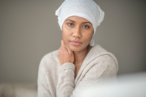 A beautiful mixed race woman sits in the comfort of her own home as she poses for a portrait.  She is dressed comfortably in a neutral sweater and a white headscarf as she looks to the camera with a neutral expression.