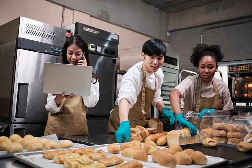 Three young friends and startup partners of bread dough and pastry foods busy with homemade baking jobs while cooking orders online, packing, and delivering on bakery shop, small business entrepreneur.