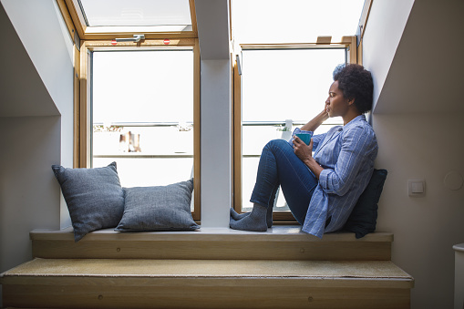 Wide shot of distraught mid adult woman sitting on the window sill, holding a cup of coffee and struggling with mental health and negative thoughts.