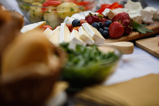 Selective focus shot of a charcuterie board with various cheese, blueberries and strawberries served on a dining table for a dinner party, among other food.