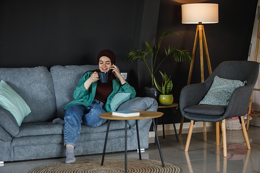 Wide shot of happy young woman, wearing a hijab, lounging on the sofa in her living room, enjoying a cup of coffee and talking on the phone with a friend.