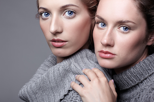Two young women in gray sweaters on grey studio background. Beautiful girls stretching hands forward in embrace. Female friendship concept.
