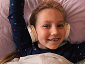 Blonde child girl in headphones learning language listening to music podcast with smartphone online in bed at home.