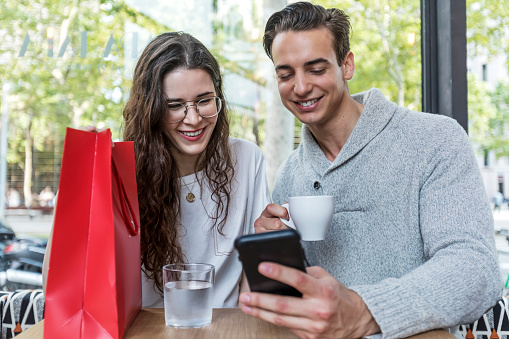 Young cheerful man and woman dating and spending time together in cafe, using phone.