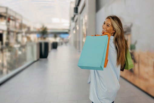 Beautiful young woman is at shopping mall, holding colorful shopping  bags over her left shoulder