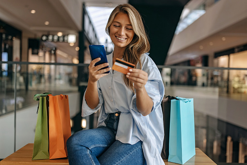 Beautiful smiling women sitting in shopping mall while holding a credit card in one hand and smart phone in the other