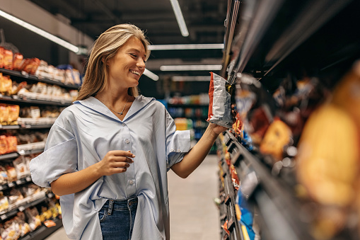 Happy young woman looking at product at grocery store. Smiling woman shopping in supermarket and using cellphone. Costumer buying food at the market.