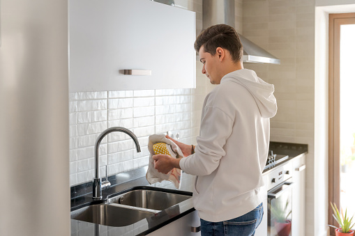 Caucasian brown-haired man in the kitchen. He is drying the dishes. In a white kitchen with black marble. There is natural light inside in Spain.