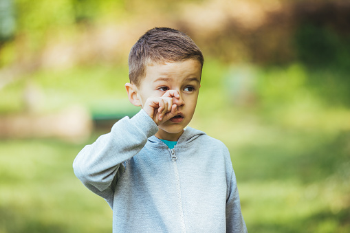 Boy rubbing eye. Child with pollen allergy. Boy sneezing because of seasonal allergy while sitting in a grass. Spring allergy concept. Flowering bushes and trees in background. Child allergy