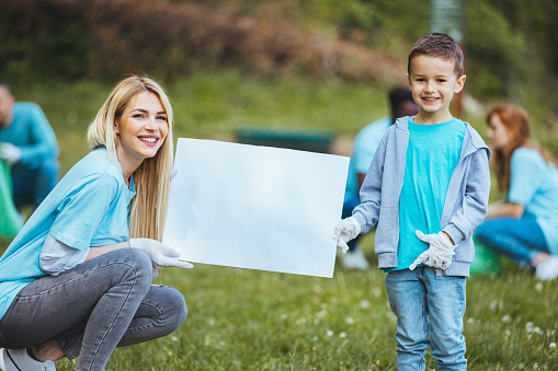 A beautiful preschool boy and his mother are standing outdoors in a public park. In the background are adults collecting garbage while they hold a sign and smile at the camera.