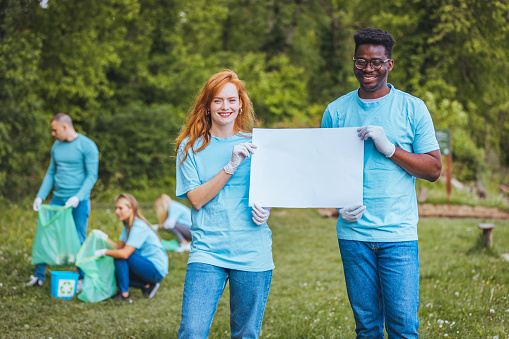 Two young people of different races work as volunteers and help clean up nature. They hold a blank banner or poster in their hands and show it directly to the camera. They look at the camera with a smile