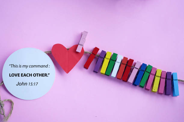 Bible verse on a paper note - This is my command, love each other. John 15:17 With red heart and colorful wooden paper clips hanging on natural rope on a purple background.