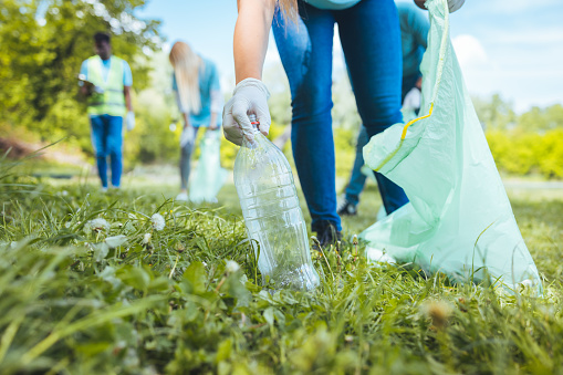 An unrecognizable woman holds a plastic garbage bottle that she puts in a recycling bag for cleaning. She is in focus. Volunteering concept