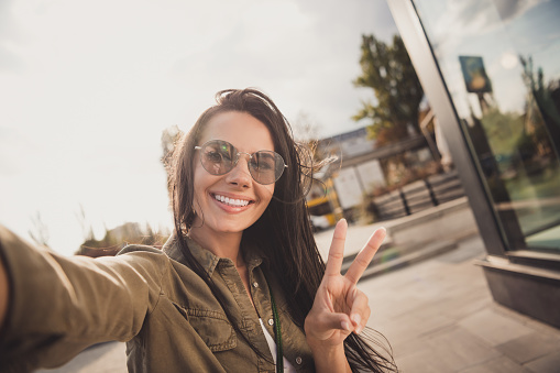 Photo of adorable sweet young lady wear brown shirt dark glasses smiling making selfie showing v-sign outside sunny street.