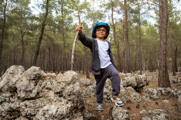 5-6 years old brunette boy with glasses. He is looking at camera in nature.\nConfident, positive emotion. Using stick in hand like trekking pole.model full body