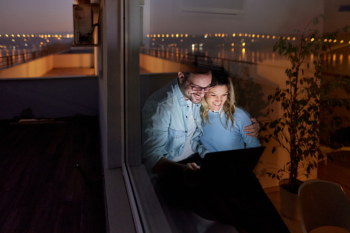 Young happy couple using computer while relaxing by the window at night. The view is through glass.