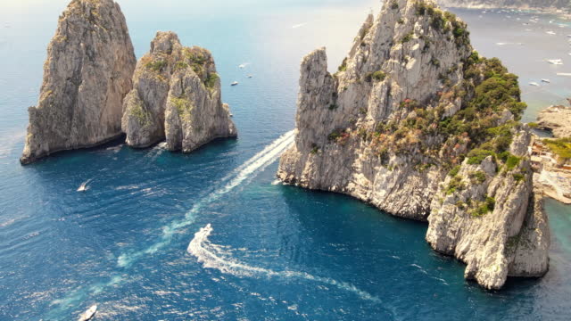 Aerial drone view of the Tyrrhenian sea coast of Capri, Italy. Rocky cliffs, blue water, floating boats, greenery