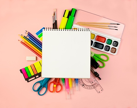 Office items on a light pink background with a white notebook in the center and a copy space. The concept of education and development. Paints ,, pencils, soda, ruler, scissors, etc. On a light banner