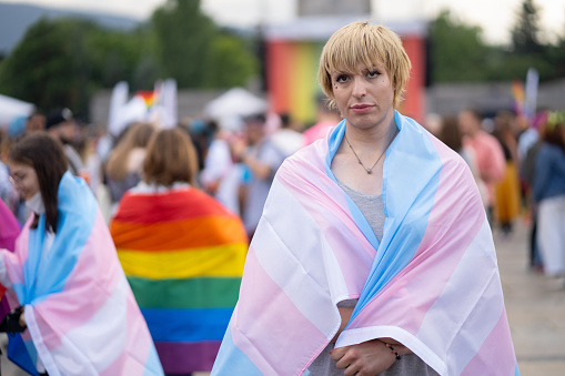 A gender-diverse person  wearing a pansexaul flag, standing in front of a crowd at a gay pride event campaigning for the rights of the LGBT+ community
