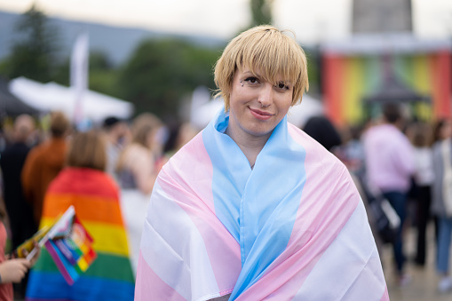 A smiling non-binary person is wrapped in a transgender flag and looking at the camera, behind are a crowd at a gay pride parade