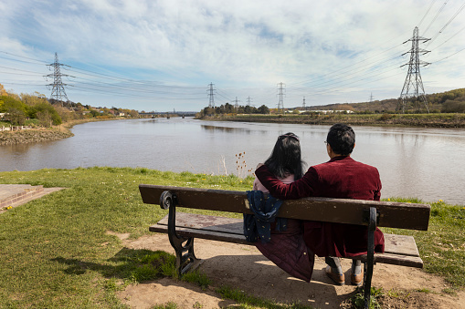 The senior male has his arm around his wife sitting on a bench riverside looking at the scenery down the river.