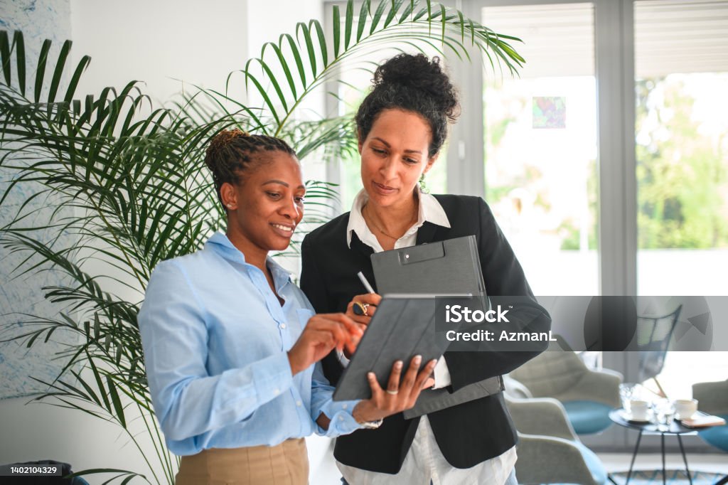 Mature Black Businesswoman And Hispanic Female Manager Working With A Digital Tablet African American entrepreneur and Hispanic female colleague working with a digital tablet at the office. They are smiling while editing online documents. Vanity Stock Photo