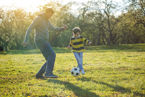 Boy playing soccer with father at public park at sunset