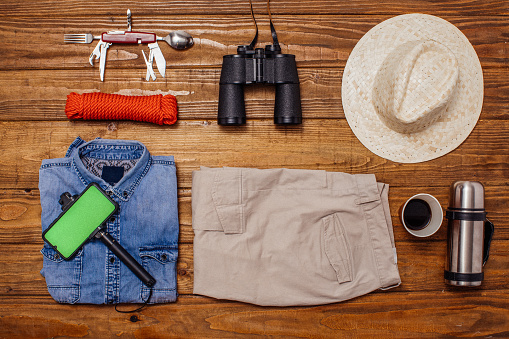 Shirt, binoculars, thermos, hat, water bottle, sunglasses and multifunctional knife on wooden background captured from above