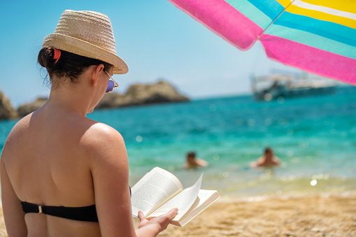 Back view of woman mid 30s, reading a book on the beach, under the parasol. during her low budget holiday. We can see hints of two tourists enjoying the water, and on horizon some rock formations and a ship.