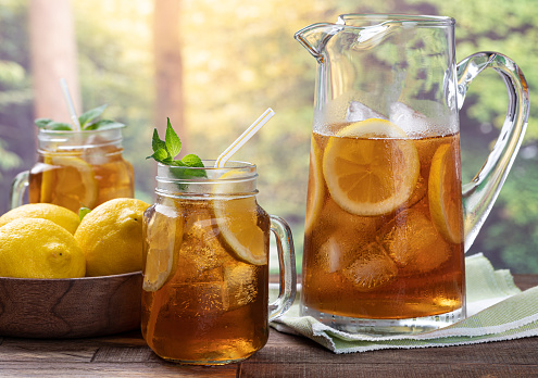 Glass and pitcher of iced tea with mint, lemon slices and ice on an outdoor wooden table with rural summer background